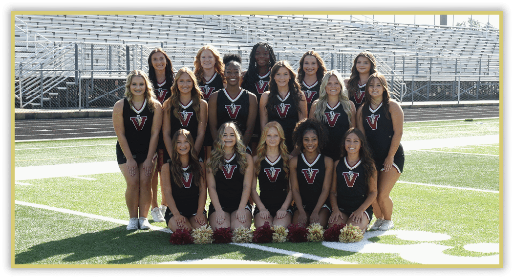 A group of people wearing matching black athletic uniforms with a red and white “V" logo pose together on a sports field with bleachers in the background. Description generated by AI