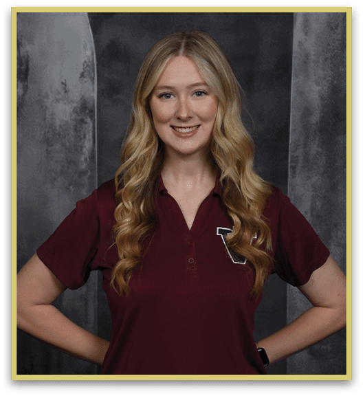A person with long wavy hair wearing a maroon polo shirt is smiling with hands on hips against a dark textured background. Description generated by AI