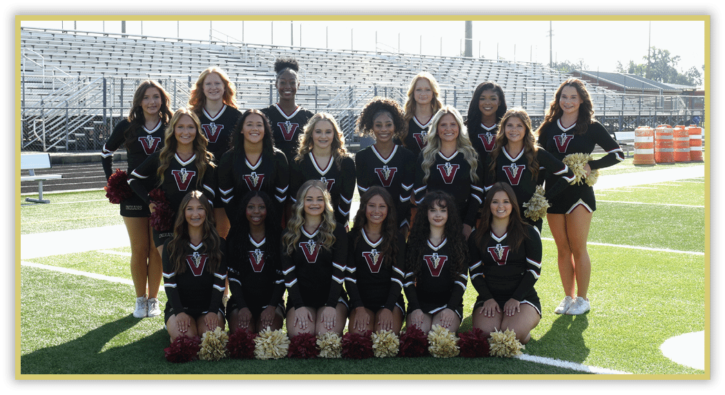 A group of adults in matching black cheerleading uniforms with a red and white logo pose on a football field with pom-poms arranged in front of them. Description generated by AI