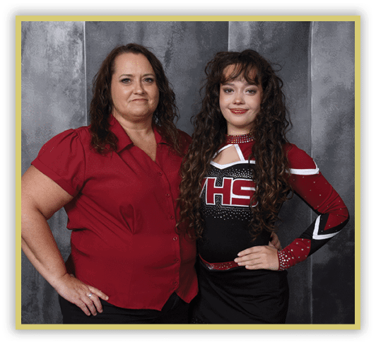 Two people stand side by side against a gray textured backdrop, one wearing a red button-up shirt and the other dressed in a black and red cheerleading uniform with long curly hair. Description generated by AI