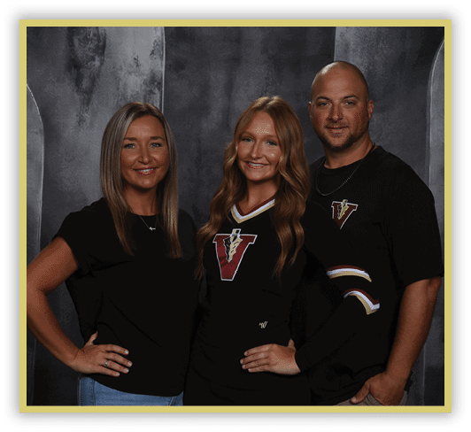 Three adults stand closely together smiling against a dark textured backdrop, with one wearing a black outfit featuring a large letter V and the others in casual black tops. Description generated by AI