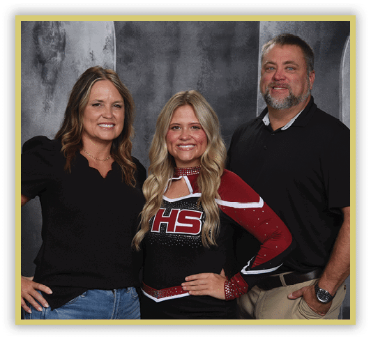 Three adults stand closely together smiling, with one wearing a cheerleading uniform and the other two dressed in casual clothing against a dark backdrop. Description generated by AI