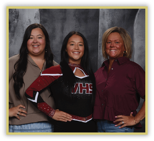 Three people stand closely together smiling, with one wearing a cheerleading uniform and the others dressed casually against a dark textured backdrop. Description generated by AI