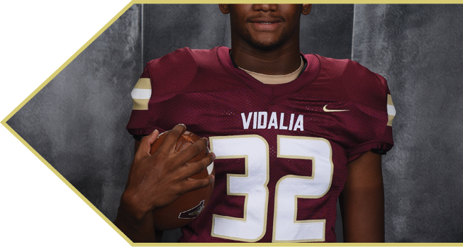 Person wearing a maroon football jersey with the number 32 and the word Vidalia holding a football against a gray background. Description generated by AI
