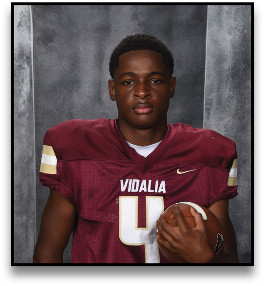 A person wearing a maroon football jersey with the number 4 and beige pants is holding a football and posing in front of a dark, textured backdrop. Description generated by AI