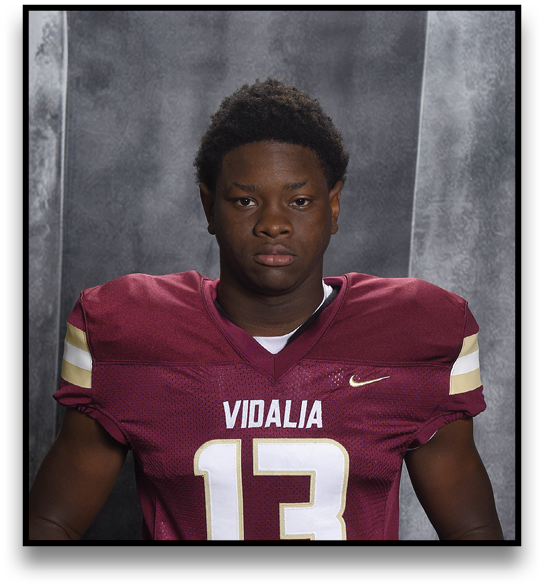 A person wearing a maroon football jersey with the number 13 and the word Vidalia is holding a football against a gray textured backdrop. Description generated by AI