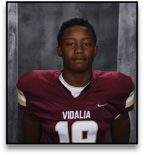 A person wearing a maroon football jersey with the number 18 and the word Vidalia stands against a dark textured background with arms behind their back. Description generated by AI