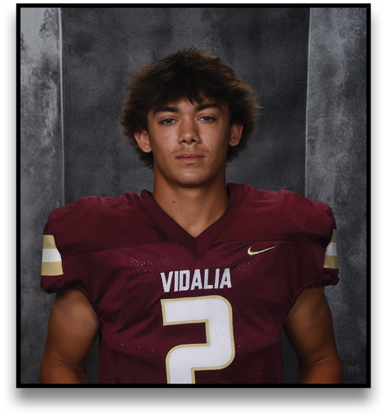 A person with tousled hair wearing a maroon football jersey with the number 2 and the word Vidalia is holding a football in front of a dark textured background. Description generated by AI