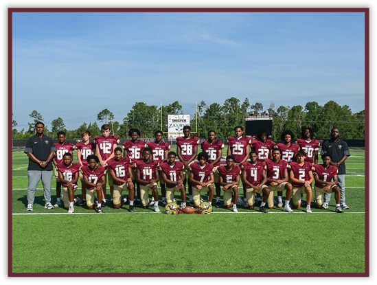 A group of people wearing maroon and gold football uniforms pose for a team photo on a green football field with two coaches standing on either side. Description generated by AI