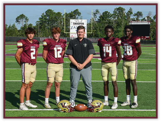 Five people wearing football uniforms and one person in casual sportswear stand on a football field with helmets and a football placed on the ground in front of them. Description generated by AI