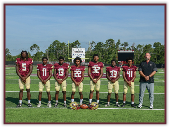 Seven people wearing matching football uniforms stand in a line on a football field with helmets and a football placed on the ground in front of them and a scoreboard visible in the background. Description generated by AI