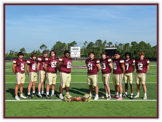 Ten people wearing maroon and gold football uniforms stand in a line on a football field with helmets and a football placed on the ground in front of them. Description generated by AI