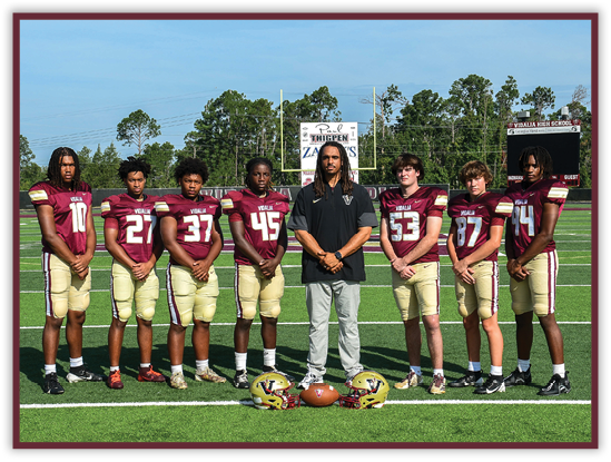 Seven people wearing matching football uniforms stand on a football field with one person in casual clothing standing in the center, and two football helmets and a football placed on the ground in front of them. Description generated by AI
