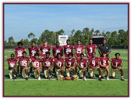 A group of people wearing maroon and gold football uniforms pose together on a football field with trees and a scoreboard in the background. Description generated by AI