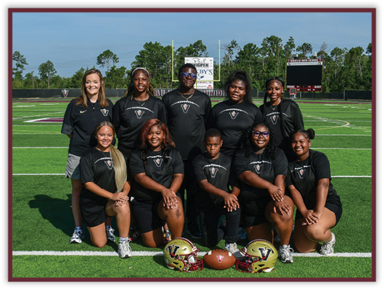 A group of adults and one child wearing matching black sports shirts pose on a football field with two helmets and a football placed in front of them. Description generated by AI