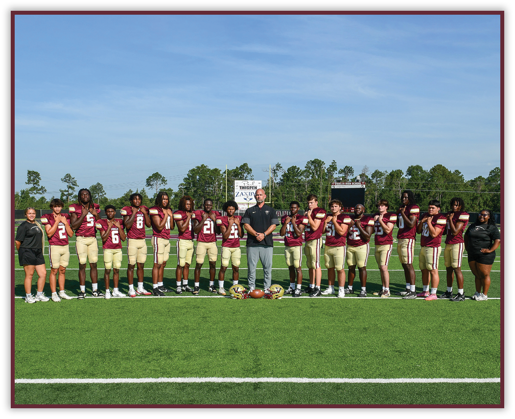 A group of people wearing matching sports uniforms and two adults stand in a line on a football field with helmets and a football placed on the ground in front of them. Description generated by AI