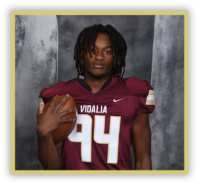 Person with dreadlocks wearing a maroon football jersey holding a football against a gray textured background. Description generated by AI