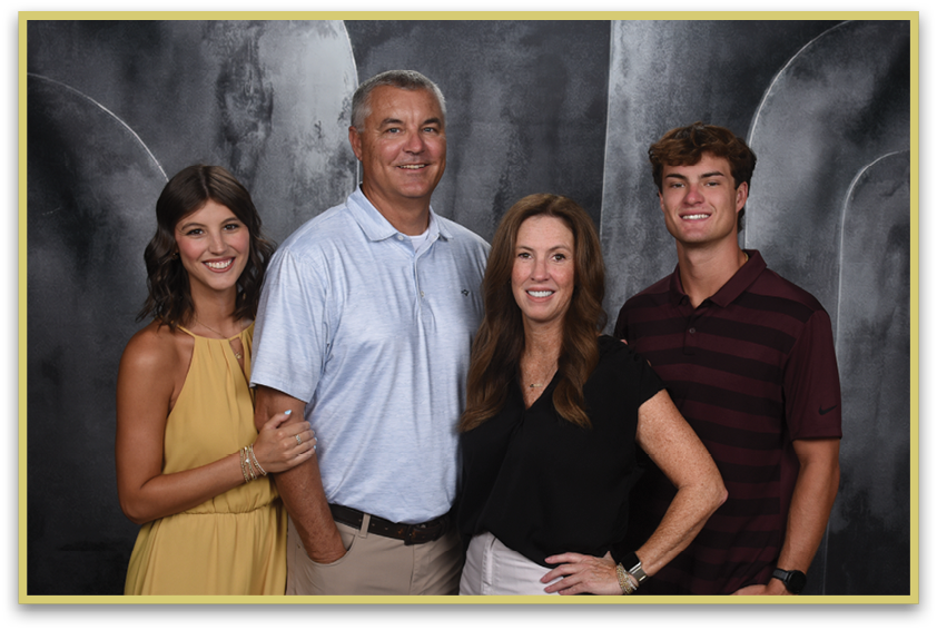 Four adults stand closely together smiling against a dark textured backdrop, with one wearing a yellow dress, one in a light blue polo shirt, one in a black top, and one in a dark striped polo shirt. Description generated by AI