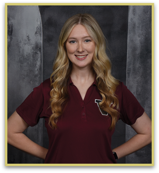 A person with long wavy hair wearing a maroon polo shirt is smiling with hands on hips against a dark textured background. Description generated by AI
