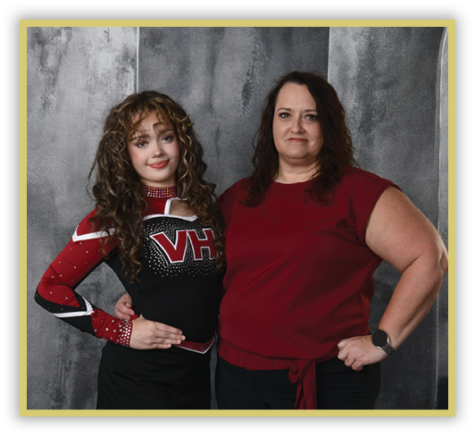 Two adults stand side by side against a gray textured backdrop, one wearing a red and black cheerleading outfit and the other wearing a red sleeveless top with a watch on their wrist. Description generated by AI