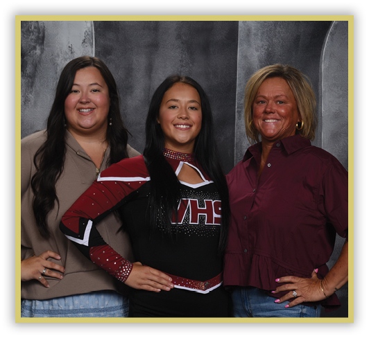 Three people stand closely together smiling, with one wearing a cheerleading uniform and the others dressed casually against a dark textured backdrop. Description generated by AI