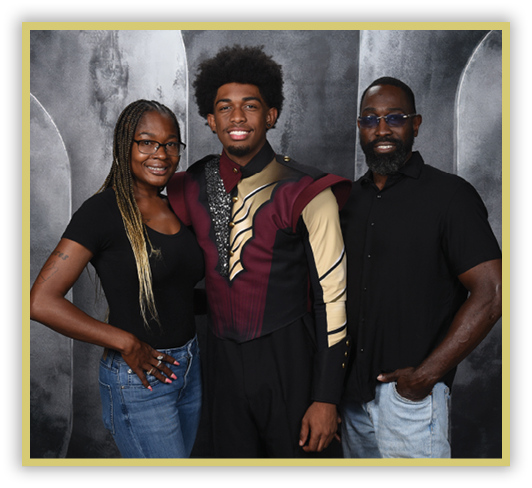 Three adults stand closely together smiling, with one wearing a decorative maroon and gold uniform, and the others dressed casually against a gray textured backdrop. Description generated by AI