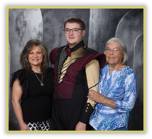Three people stand closely together against a gray backdrop, with one person in the center wearing a maroon and black uniform with gold accents, while the others wear casual clothing and smile. Description generated by AI