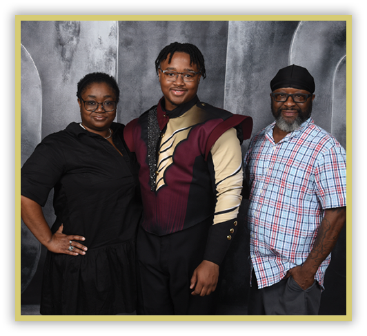 Three adults stand closely together smiling, with one wearing a maroon and beige uniform, another in a black dress, and the third in a plaid shirt and head covering against a gray textured backdrop. Description generated by AI