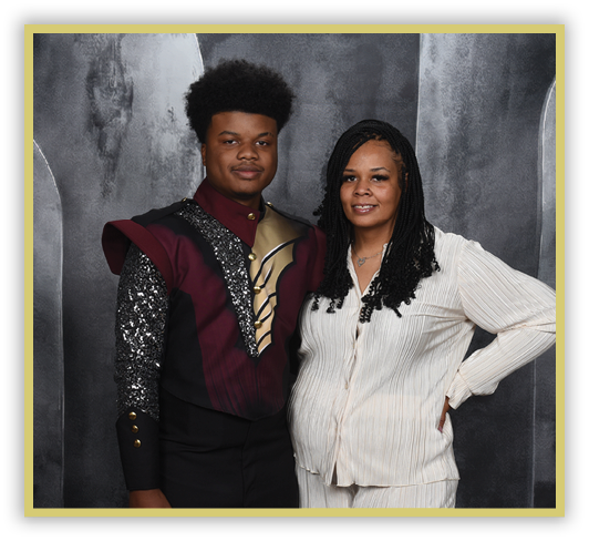 Two people stand side by side against a dark textured backdrop, one wearing a decorative uniform with sequins and gold accents and the other dressed in a light-colored striped outfit with long braided hair. Description generated by AI