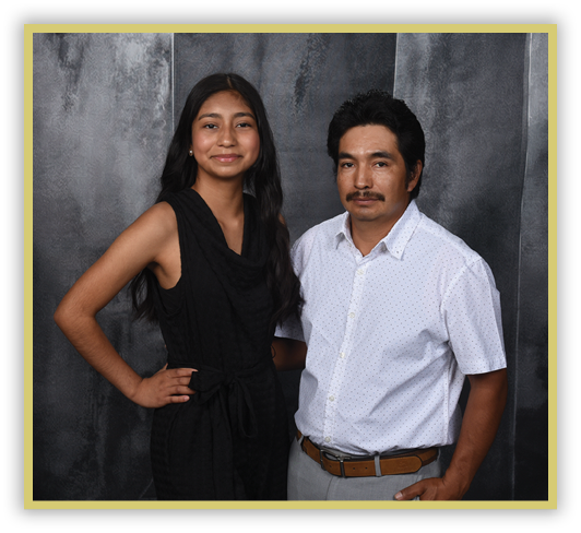 Two adults stand side by side against a dark textured background, one wearing a sleeveless black outfit and the other wearing a white short-sleeve button-up shirt with light-colored pants and a brown belt. Description generated by AI