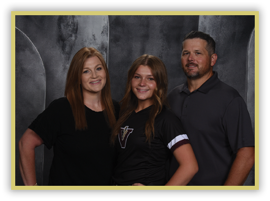 Three adults stand closely together smiling against a dark textured backdrop, with one wearing a black shirt with a logo and the others in solid dark tops. Description generated by AI