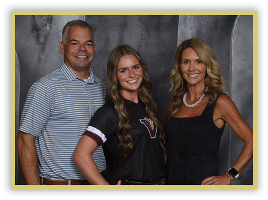 Three adults stand closely together smiling against a dark gray backdrop, with one wearing a striped polo shirt, another in a black sports jersey, and the third in a black sleeveless dress with a necklace. Description generated by AI
