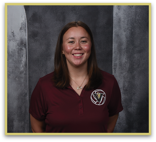 A person with shoulder-length straight hair is smiling and wearing a maroon polo shirt with a logo on the left chest against a gray textured background. Description generated by AI