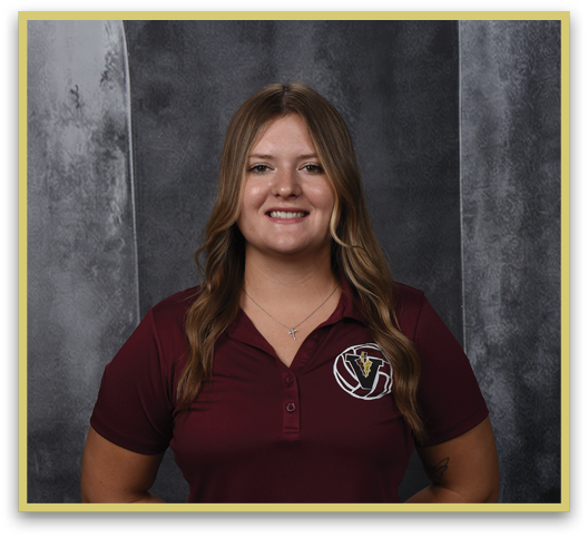 A person with long wavy hair wearing a maroon polo shirt with a logo and a cross necklace is smiling against a dark textured background. Description generated by AI