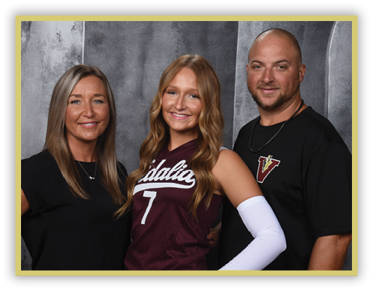 Three adults stand closely together smiling, with one wearing a sports jersey and arm sleeve, and the others dressed in casual clothing against a gray backdrop. Description generated by AI