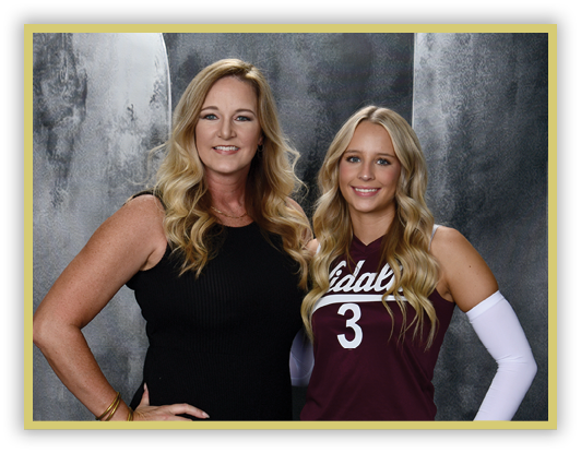 Two adults with long wavy hair stand side by side smiling, one wearing a black sleeveless dress and the other in a maroon sports jersey with the number three and a white arm sleeve, against a gray textured backdrop. Description generated by AI