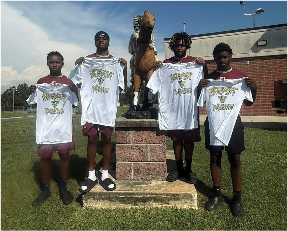 Four people standing outdoors on grass in front of a horse statue holding white shirts with text on them under a clear blue sky. Description generated by AI