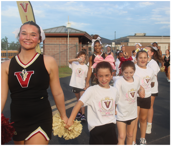 A group of children and adults dressed in cheerleading uniforms and t-shirts are standing outdoors on a paved surface, holding hands and smiling. Description generated by AI