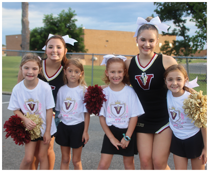 Four people wearing cheerleading outfits and white bows are standing outdoors with a fence and building in the background, two of them holding red pom-poms and smiling. Description generated by AI
