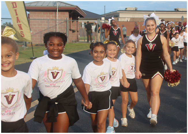 A group of children and a cheerleader in uniform are walking hand in hand on a paved path near a sports field with banners reading “Indians" in the background. Description generated by AI