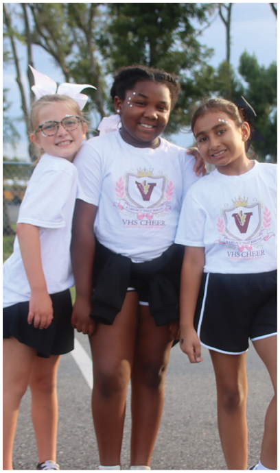 Three people wearing matching white t-shirts and black shorts stand closely together outdoors on a paved surface with a chain-link fence and trees in the background. Description generated by AI