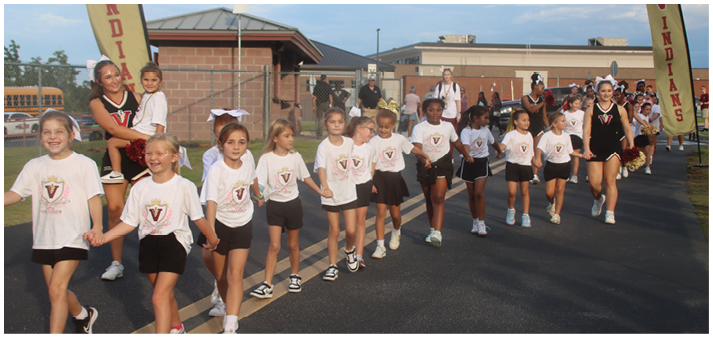 A group of children wearing matching white t-shirts and black shorts are walking hand in hand on a paved path near a school building with adults and other children in the background. Description generated by AI