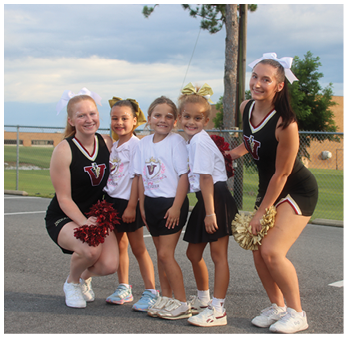 Two adults in cheerleading uniforms and three children in matching t-shirts and skirts pose together outdoors on a paved surface with a chain-link fence and trees in the background. Description generated by AI
