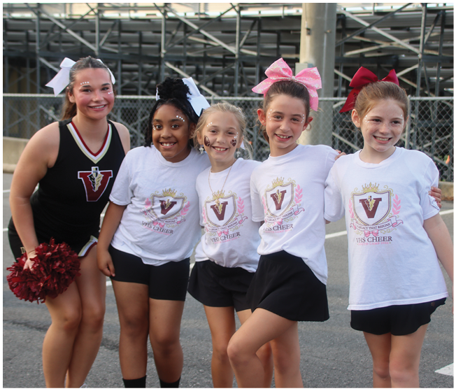 Five children wearing cheerleading outfits and bows are standing together outdoors smiling and posing for a photo. Description generated by AI
