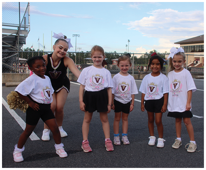 Five children wearing matching white t-shirts with a logo and black bottoms stand together outdoors on a paved surface with one child in a cheerleading outfit and bow posing with them. Description generated by AI