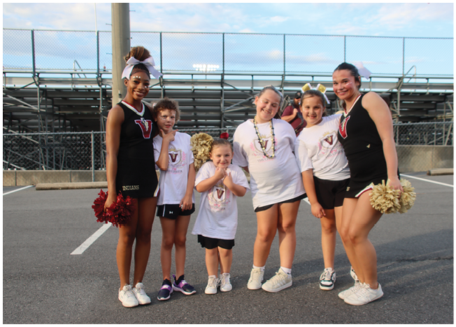 A group of six people wearing cheerleading outfits and casual sportswear stand together smiling in an outdoor parking lot near bleachers. Description generated by AI