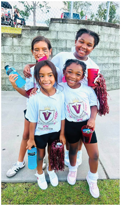 Four children wearing white shirts smile and pose closely together outdoors in front of a stone wall and chain-link fence. Description generated by AI