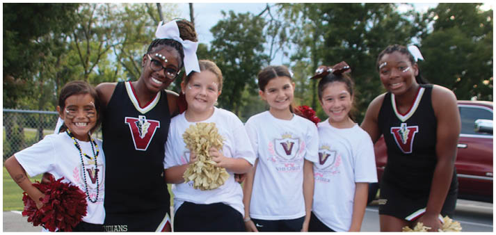 A group of six children wearing cheerleading outfits and holding pom-poms stand together outdoors on a paved surface with trees and a parked vehicle in the background. Description generated by AI