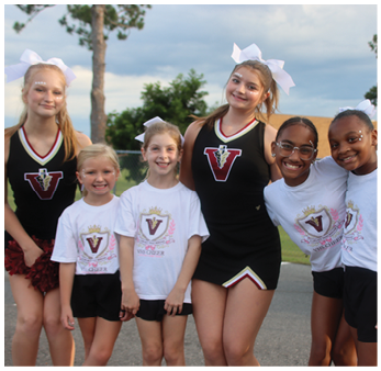 A group of six children wearing cheerleading uniforms and white bows are standing outdoors on a paved surface with trees and grass in the background. Description generated by AI