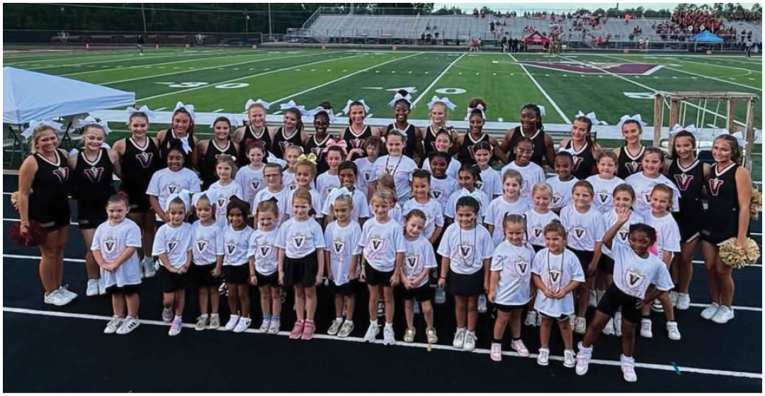 A large group of children and adults wearing matching cheerleading uniforms pose together on a track in front of a football field under a clear sky. Description generated by AI